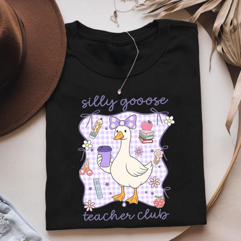 May include: Black t-shirt featuring a cartoon goose holding a purple coffee cup, with the text "silly goooose teacher club." The design includes a purple bow, pencils, books, and flowers. The goose is standing on a purple and white checkered background.