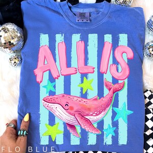 以下が含まれることがあります： ピンクのクジラのイラスト、ピンクのブロック文字で「ALLIS」という名前、緑と青の星が描かれたフロブルーのTシャツ。Tシャツはクルーネックと半袖です。テキスト「FLO BLUE」はシャツの下部にあります。