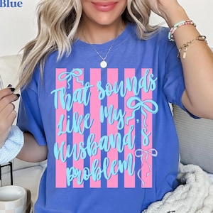 May include: Blue t-shirt with the text "That sounds like my husband's problem" in light blue script. The text is set against a pink striped background with bow accents. The word "Blue" is in the top left corner.