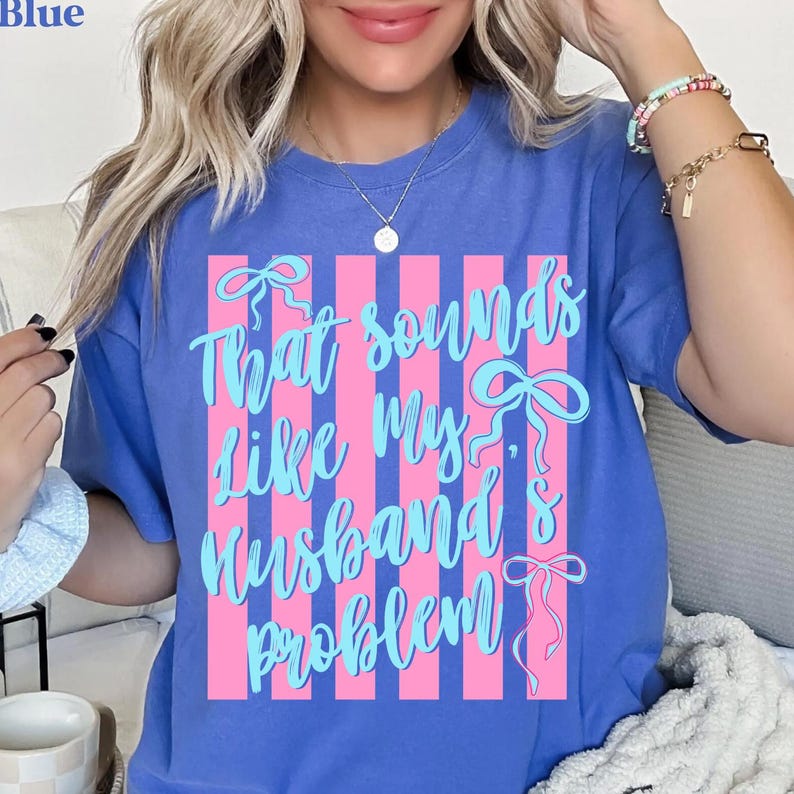 May include: Blue t-shirt with the text "That sounds like my husband's problem" in light blue script. The text is set against a pink striped background with bow accents. The word "Blue" is in the top left corner.