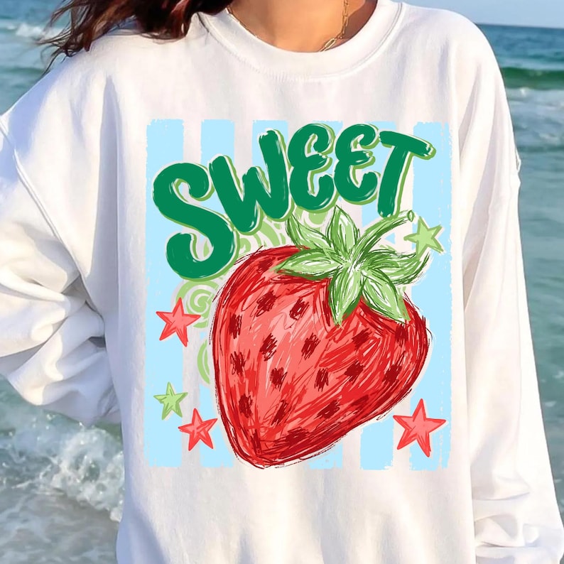 May include: White sweatshirt featuring a large red strawberry with green leaves and the word "SWEET" in green script. The design includes red and green stars on a light blue striped background. A casual, comfortable top.