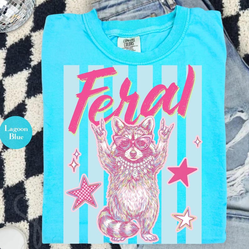 Pode incluir: Camiseta azul lagoa com um gr&aacute;fico de guaxinim usando &oacute;culos de sol e um colar, com a palavra "Feral" em rosa. O guaxinim est&aacute; fazendo um gesto de rock and roll com a m&atilde;o. Estrelas rosas e uma estrela xadrez tamb&eacute;m est&atilde;o na camiseta.