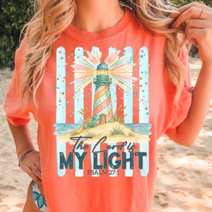 Pu&ograve; includere: T-shirt color corallo con un design a faro colorato. Il faro ha un motivo a strisce e si trova su una piccola isola con il testo "The Lord is My Light" e Salmo 27:1.