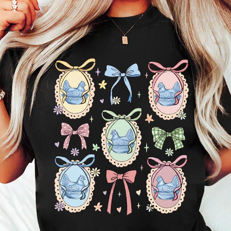 May include: Black t-shirt featuring a repeating pattern of oval frames with a blue chicken illustration, adorned with colorful bows and floral accents. The design includes pink, yellow, blue, and green elements, creating a whimsical and decorative aesthetic.