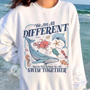 May include: White sweatshirt with a colorful graphic of various sea creatures, including a whale, octopus, and dolphin. The text on the sweatshirt reads "We Are All DIFFERENT" and "But In This School We All SWIM TOGETHER."
