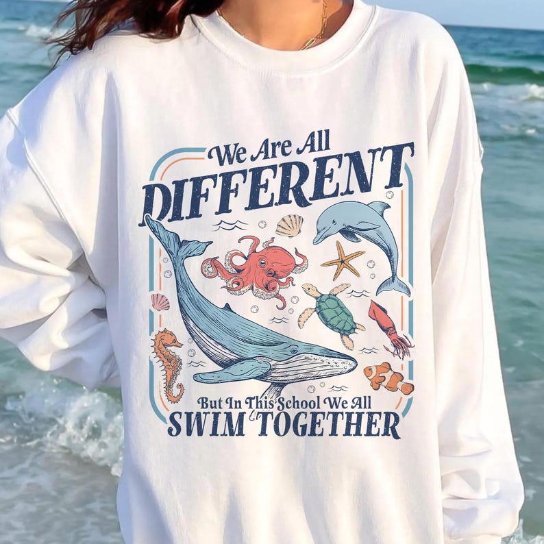 May include: White sweatshirt with a colorful graphic of various sea creatures, including a whale, octopus, and dolphin. The text on the sweatshirt reads "We Are All DIFFERENT" and "But In This School We All SWIM TOGETHER."