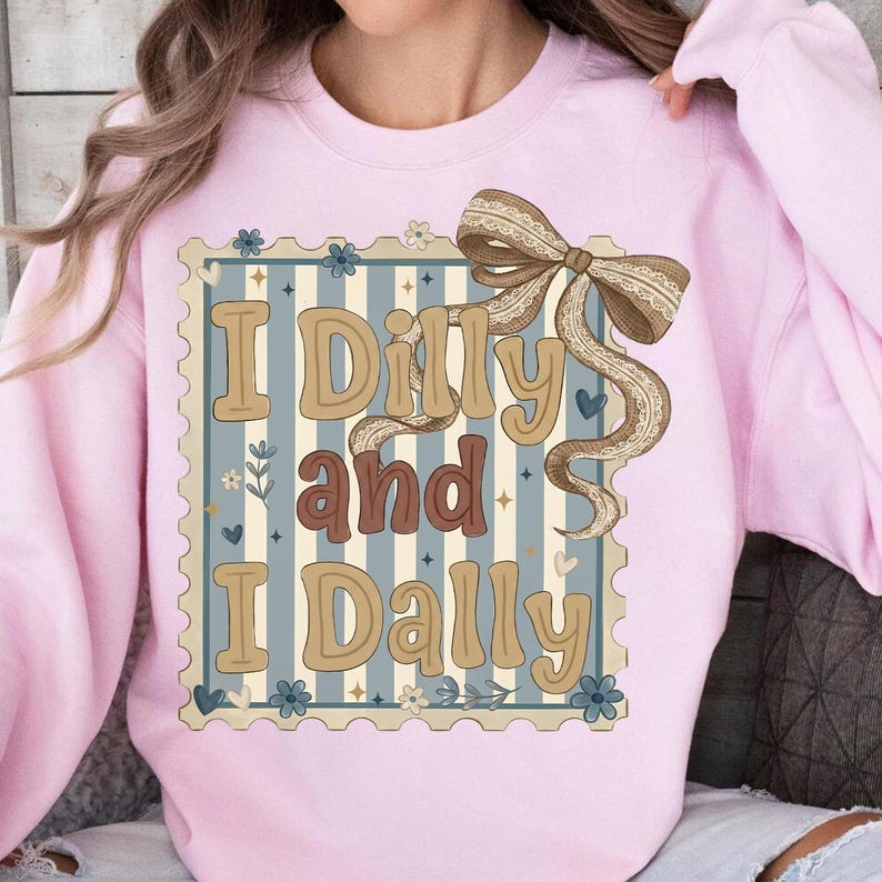 May include: A light pink sweatshirt featuring a decorative design with the text "I Dilly and I Dally". The design includes a postage stamp-like border, a striped background, and a brown bow.