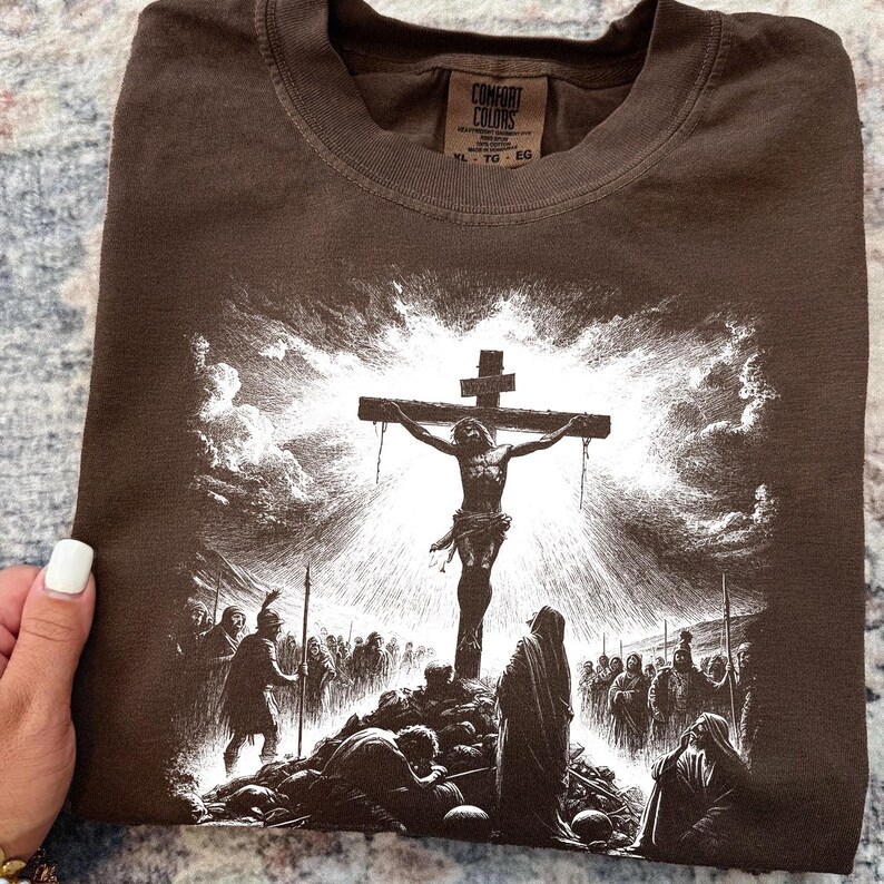 May include: Brown t-shirt featuring a detailed black and white illustration of the crucifixion of Jesus Christ. The graphic depicts Jesus on the cross, surrounded by figures and a dramatic sky. The shirt has a crew neck and a tag that says "Comfort Colors".