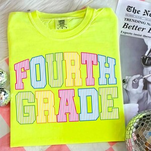 May include: A neon yellow short-sleeve t-shirt featuring the text "FOURTH GRADE" in a collegiate font. The letters are filled with vertical stripes in alternating colors of pink, blue, yellow, and green. A Comfort Colors brand tag is at the neck.