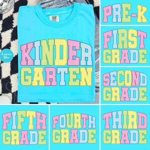 May include: A lagoon blue t-shirt featuring the word KINDERGARTEN in large, block-style letters. Each letter has a vertical striped pattern in alternating pastel colors including pink, yellow, and light blue. The shirt has a crew neckline.