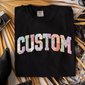 May include: Black crew neck sweatshirt with the word "CUSTOM" in patchwork letters. The letters are made of various floral and gingham fabrics in pastel colors. The sweatshirt has a tag that says "COMFORT COLORS" and the size is XL-TG-EG.