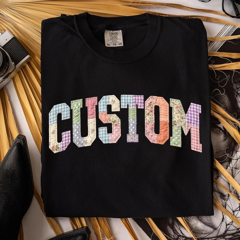 May include: Black crew neck sweatshirt with the word "CUSTOM" in patchwork letters. The letters are made of various floral and gingham fabrics in pastel colors. The sweatshirt has a tag that says "COMFORT COLORS" and the size is XL-TG-EG.