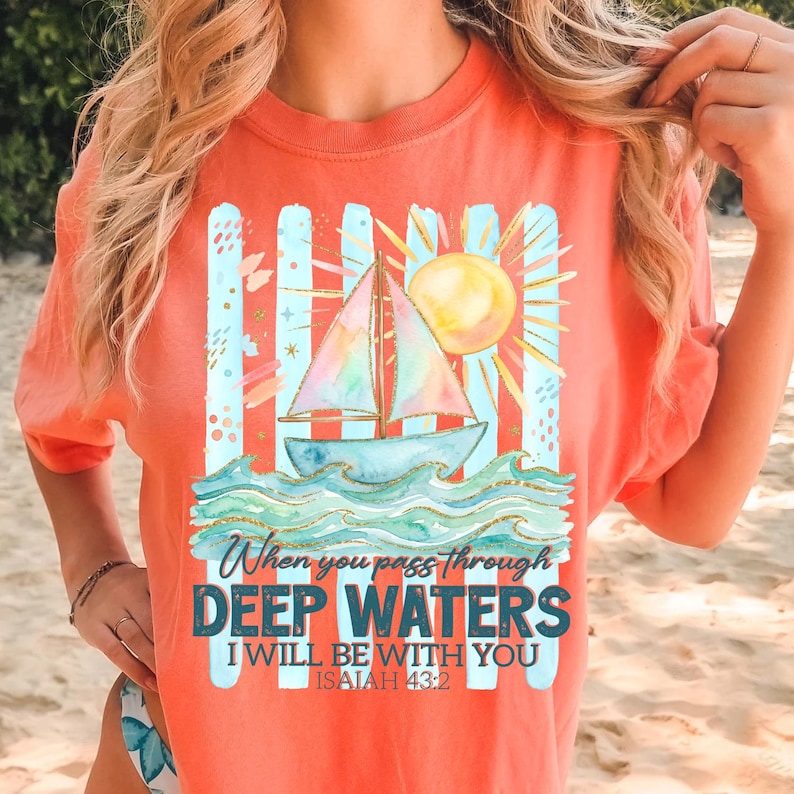 K&ouml;nnte beinhalten: Korallfarbenes T-Shirt mit einem Segelboot-Motiv, Sonne und Wellen. Der Aufdruck enth&auml;lt den Text "When you pass through Deep Waters, I will be with you, Isaiah 43:2". Das Shirt hat einen Rundhalsausschnitt und kurze &Auml;rmel.