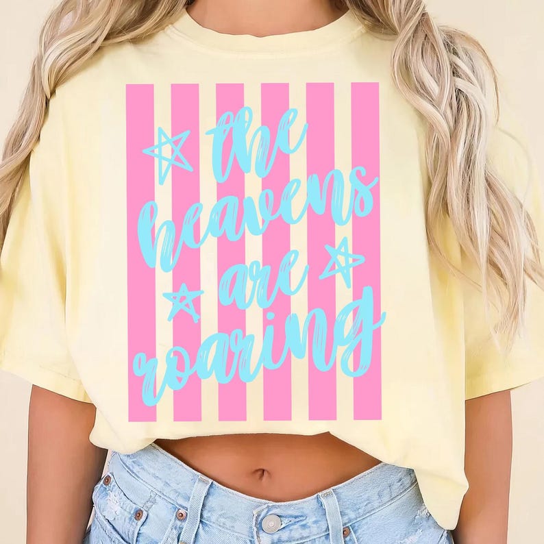 May include: A light yellow t-shirt with a pink and white striped design. The shirt features the text "the heavens are roaring" in a light blue script font with star accents. The shirt is made of a soft material.