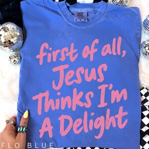 May include: A blue t-shirt with the text "first of all, Jesus Thinks I'm A Delight" in pink. The shirt is a Comfort Colors brand and is a medium size. The shirt is on a white surface with disco balls.