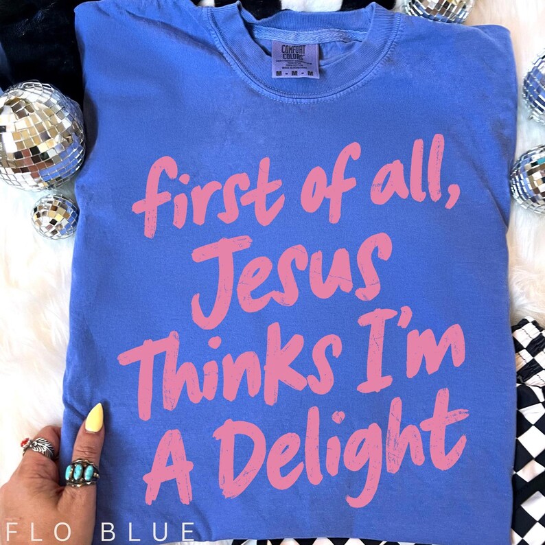May include: A blue t-shirt with the text "first of all, Jesus Thinks I'm A Delight" in pink. The shirt is a Comfort Colors brand and is a medium size. The shirt is on a white surface with disco balls.