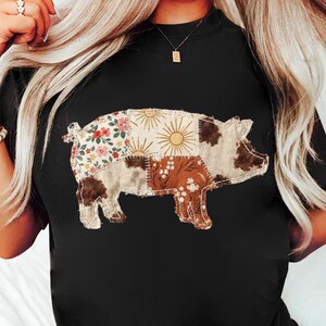 May include: Black t-shirt featuring a patchwork pig design. The pig is composed of various fabric patterns, including floral, sunburst, and cowhide prints. The design is centered on the shirt, creating a rustic and country-inspired aesthetic.