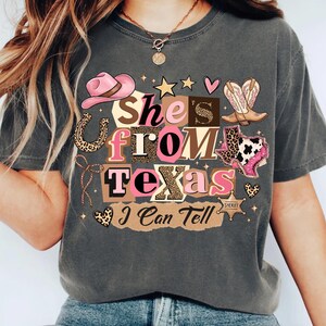 May include: Dark gray t-shirt with the text "She's From Texas I Can Tell" in a collage of pink, brown, and leopard print letters and graphics. The design includes a pink cowboy hat, boots, and a Texas outline.