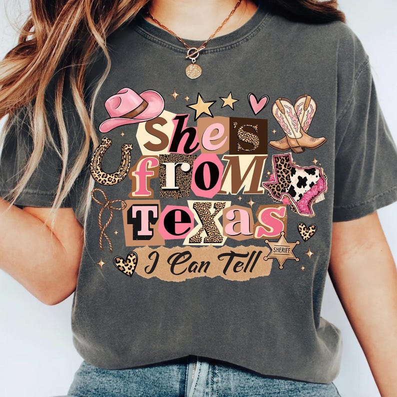 May include: Dark gray t-shirt with the text "She's From Texas I Can Tell" in a collage of pink, brown, and leopard print letters and graphics. The design includes a pink cowboy hat, boots, and a Texas outline.