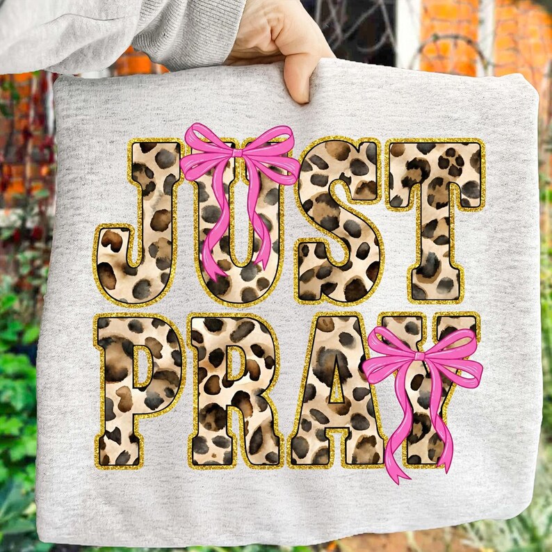 May include: A light gray sweatshirt featuring the words "JUST PRAY" in a leopard print design with gold glitter outlines. Pink bows are placed on the letters "J", "U", and "Y".