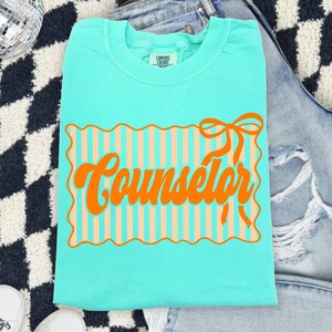 May include: A turquoise t-shirt with the word "Counselor" in orange script. The word is inside a rectangular design with peach-colored vertical stripes and a wavy border. A bow is above the word.