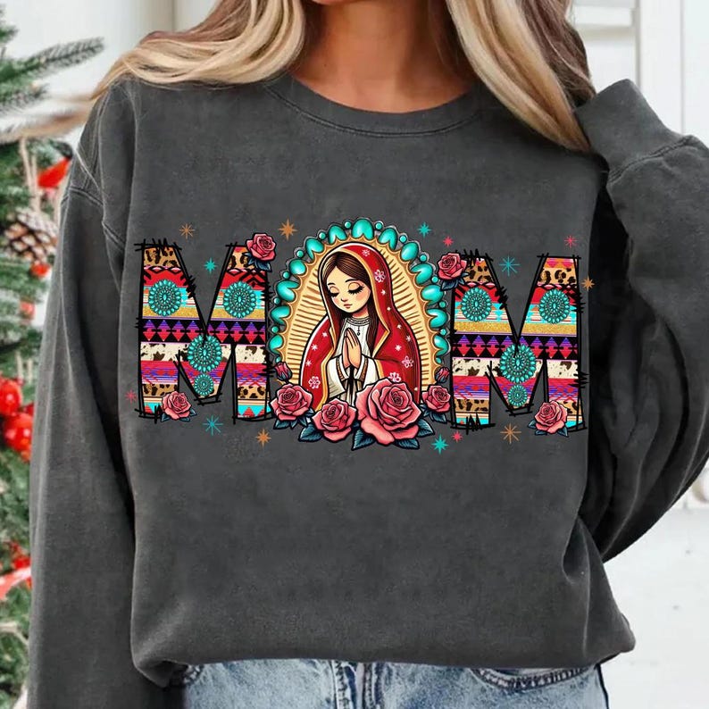 May include: Dark gray sweatshirt featuring the word "MOM" in a colorful, patterned design with floral accents. The design includes an illustration of a religious figure with a red shawl and a halo, surrounded by pink roses and turquoise accents.