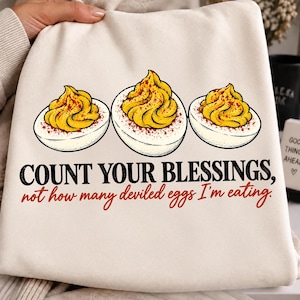 May include: A cream-colored sweatshirt featuring a graphic of three deviled eggs with yellow yolks and red seasoning. The text reads: COUNT YOUR BLESSINGS, not how many deviled eggs I'm eating. The design is centered on the front of the garment.