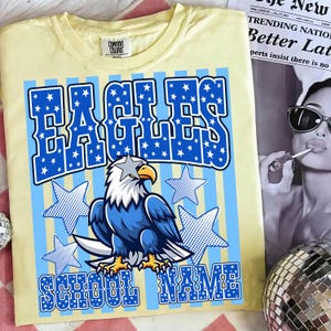 May include: A light yellow t-shirt with a blue and white graphic design. The design features the word "EAGLES" in large blue letters filled with white stars, an eagle illustration, and the words "SCHOOL NAME".