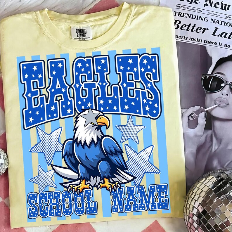 May include: A light yellow t-shirt with a blue and white graphic design. The design features the word "EAGLES" in large blue letters filled with white stars, an eagle illustration, and the words "SCHOOL NAME".