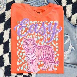 May include: Orange t-shirt with a pink tiger graphic and the word "Easy" in purple script. The tiger is set against a light blue leopard print pattern. The shirt is folded and placed on a denim background.