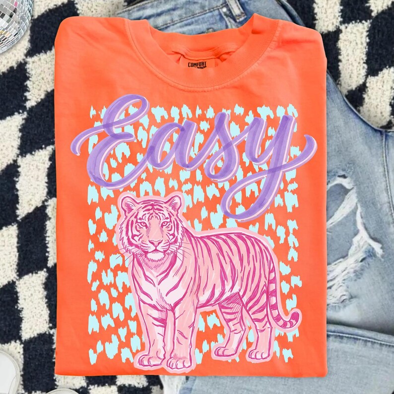May include: Orange t-shirt with a pink tiger graphic and the word "Easy" in purple script. The tiger is set against a light blue leopard print pattern. The shirt is folded and placed on a denim background.