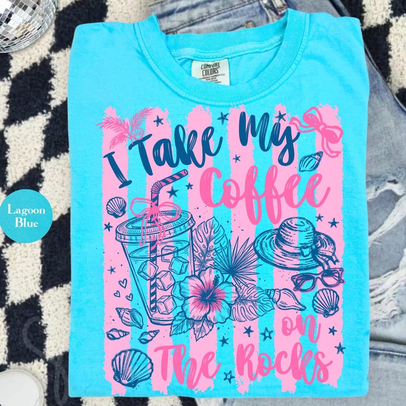 May include: Lagoon blue t-shirt with a graphic design. The design features the text "I take my coffee on the rocks" with illustrations of a coffee drink, hat, sunglasses, and seashells in pink and blue.