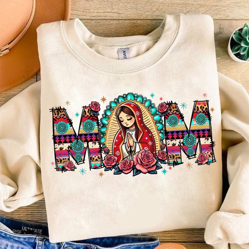 May include: Cream-colored sweatshirt with the word "MOM" in a colorful, patterned design. The letter "O" features an illustration of a religious figure with a red shawl, surrounded by roses and a turquoise halo.