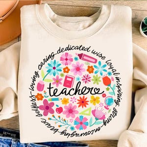May include: A white sweatshirt with a circle design featuring colorful flowers, school supplies, and the word "teachers" in the center. The circle is surrounded by a phrase that reads "caring-dedicated-wise-loyal-inspiring-strong-brave-amazing-fun-loving-perfect".