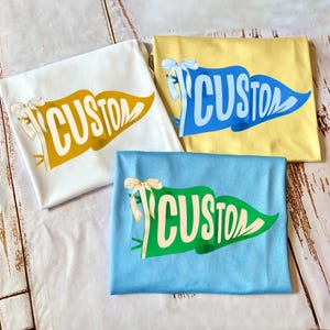 May include: Three folded t-shirts in white, yellow, and blue, each featuring a pennant design with the word "CUSTOM" in a contrasting color. Each shirt has a decorative bow.