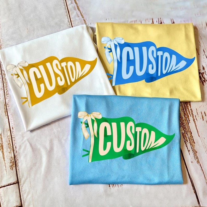 May include: Three folded t-shirts in white, yellow, and blue, each featuring a pennant design with the word "CUSTOM" in a contrasting color. Each shirt has a decorative bow.