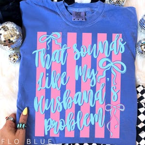 May include: Flo blue t-shirt with the text "That sounds like my husband's problem" in light blue script. The design includes pink vertical stripes and bow accents. The shirt is a Comfort Colors brand.
