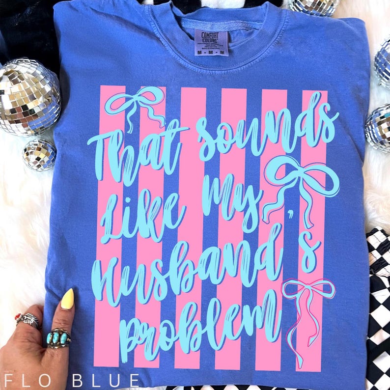 May include: Flo blue t-shirt with the text "That sounds like my husband's problem" in light blue script. The design includes pink vertical stripes and bow accents. The shirt is a Comfort Colors brand.