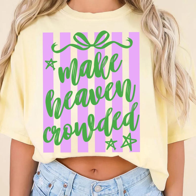 May include: A light yellow t-shirt with a graphic that says "Make Heaven Crowded" in green script. The graphic is set against a background of vertical purple and white stripes, with green stars and a bow.