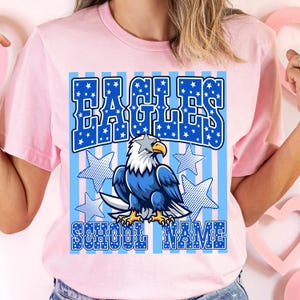 May include: Pink t-shirt featuring a blue and white graphic design. The design includes the word "EAGLES" in a blue, star-filled font, an eagle illustration, and the words "SCHOOL NAME". The eagle is blue, white, and yellow.