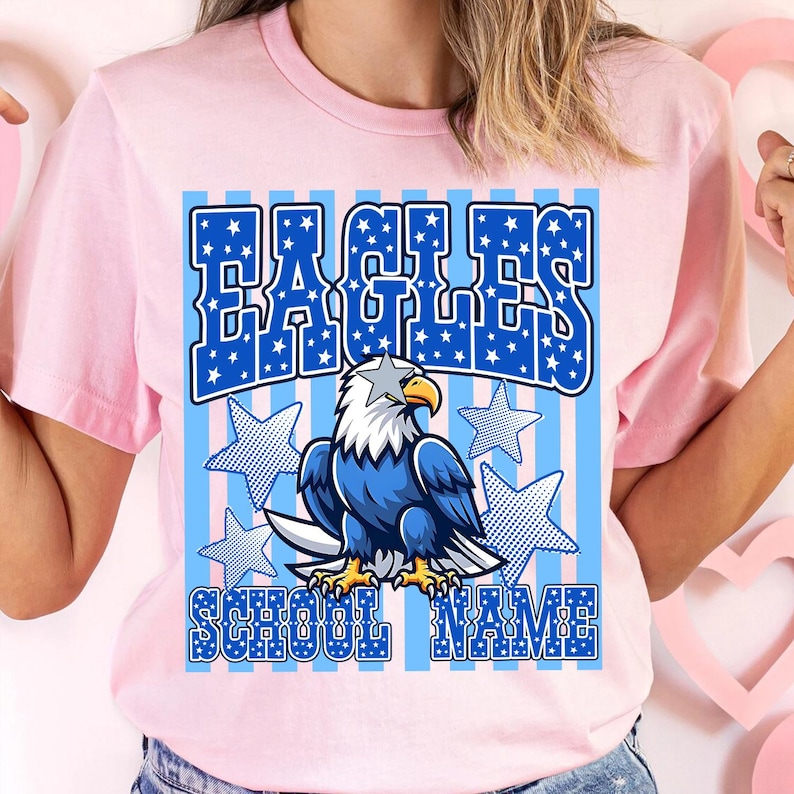 May include: Pink t-shirt featuring a blue and white graphic design. The design includes the word "EAGLES" in a blue, star-filled font, an eagle illustration, and the words "SCHOOL NAME". The eagle is blue, white, and yellow.