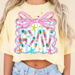 May include: A light yellow t-shirt featuring a colorful graphic design. The design includes the letters "GKV" in a striped pattern, a pink bow, stars, and a blue and white striped background. The shirt is a casual style.