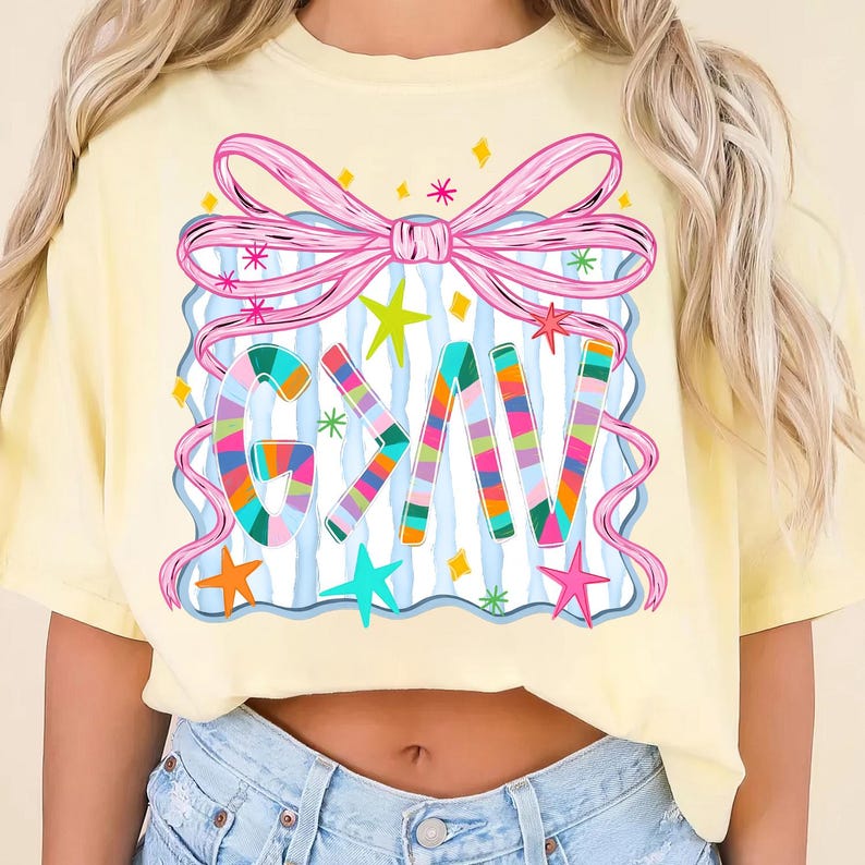 May include: A light yellow t-shirt featuring a colorful graphic design. The design includes the letters "GKV" in a striped pattern, a pink bow, stars, and a blue and white striped background. The shirt is a casual style.
