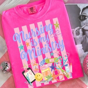May include: Bright pink crew neck t-shirt with the phrase "Nursing is my Ministry" in blue script. The design includes medical-themed illustrations like a stethoscope, scissors, and a smiley face. The shirt has a Comfort Colors tag.