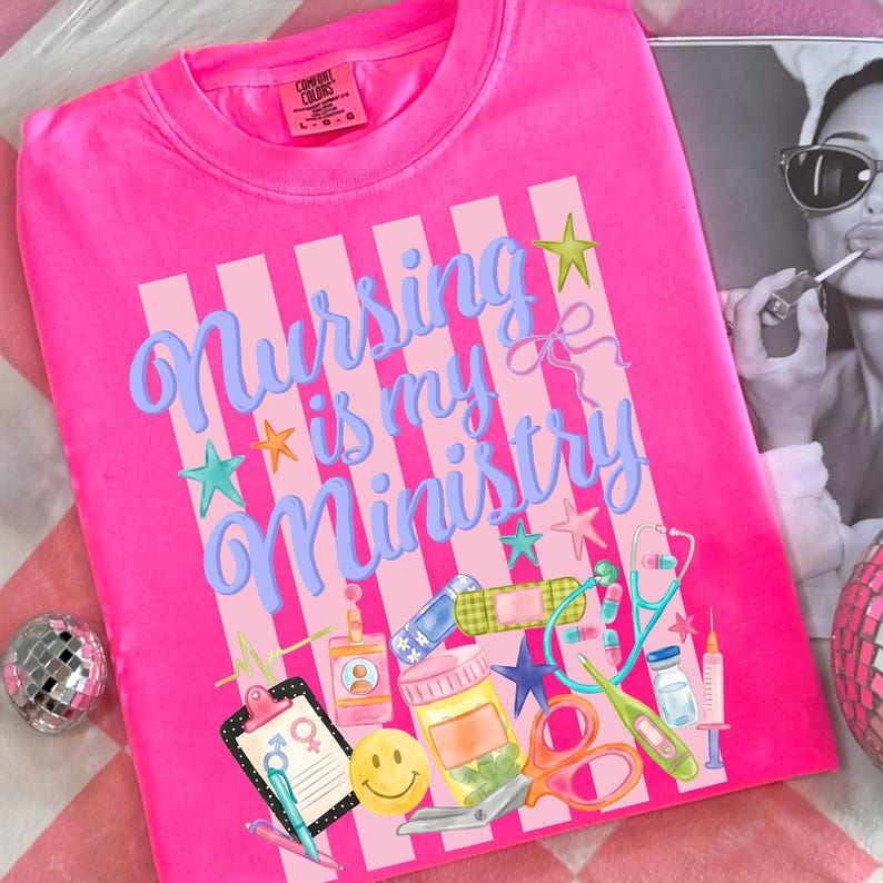 May include: Bright pink crew neck t-shirt with the phrase "Nursing is my Ministry" in blue script. The design includes medical-themed illustrations like a stethoscope, scissors, and a smiley face. The shirt has a Comfort Colors tag.