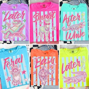 Pode incluir: Uma cole&ccedil;&atilde;o de camisetas coloridas com v&aacute;rios designs. Cada camisa apresenta um gr&aacute;fico de animal diferente, como um jacar&eacute;, um burro, um guaxinim e um tigre, junto com textos como "Later" e "Smart". As camisas t&ecirc;m um fundo listrado.