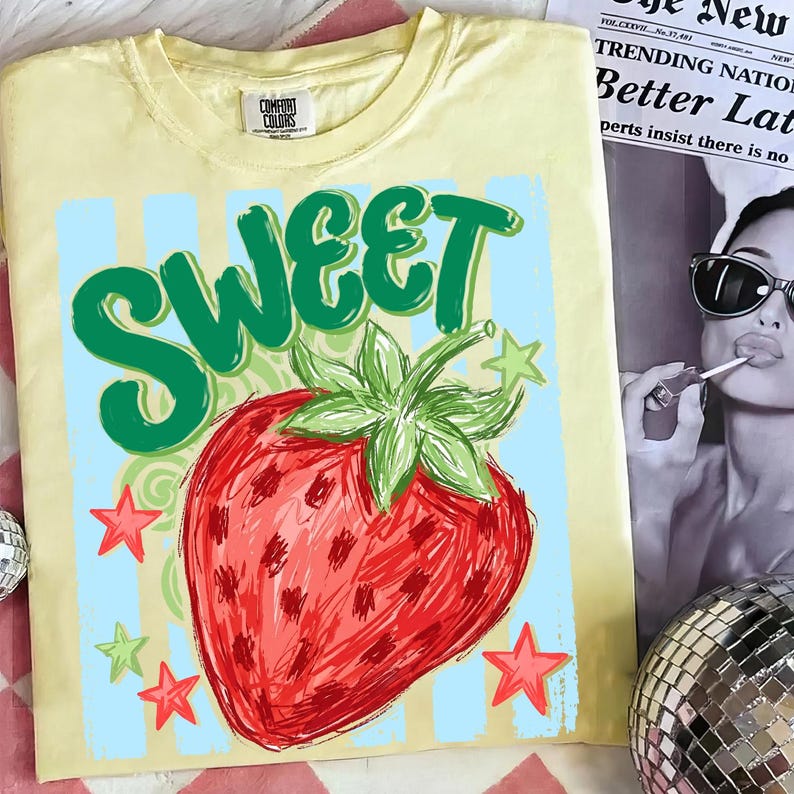 May include: Yellow t-shirt with a graphic of a red strawberry and the word "SWEET" in green. The design includes blue stripes, green stars, and red stars. The shirt has a Comfort Colors tag.