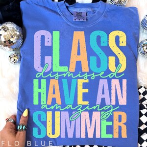 May include: Flo blue t-shirt with the words "CLASS DISMISSED HAVE AN AMAZING SUMMER" in colorful block letters. The shirt has a crew neck and a Comfort Colors tag. The text is in pastel colors.