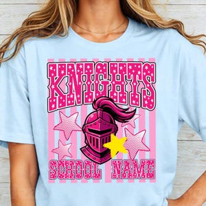 May include: Light blue t-shirt with a pink and white graphic. The graphic features the word "KNIGHTS" in pink, a pink knight's helmet, pink stars, and the words "SCHOOL NAME" in pink. The shirt is a crew neck.