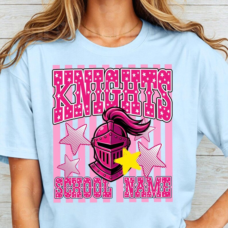 May include: Light blue t-shirt with a pink and white graphic. The graphic features the word "KNIGHTS" in pink, a pink knight's helmet, pink stars, and the words "SCHOOL NAME" in pink. The shirt is a crew neck.